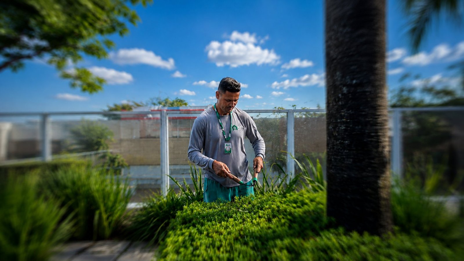 Profissional uniformizado da R2 Service aparando arbustos em jardim corporativo elevado, sob céu azul